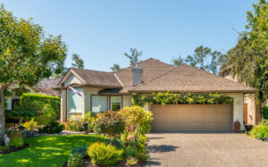 Beautiful single-story custom home in Oakhurst, California, featuring a manicured green lawn, vibrant front yard landscaping, and a beige two-car garage with a lush green vine trellis.