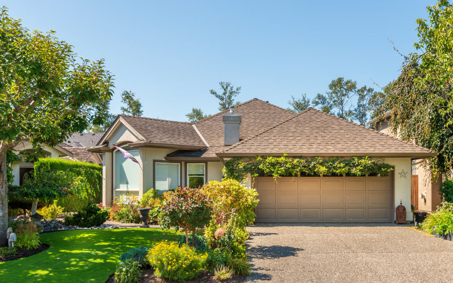 Beautiful single-story custom home in Oakhurst, California, featuring a manicured green lawn, vibrant front yard landscaping, and a beige two-car garage with a lush green vine trellis.