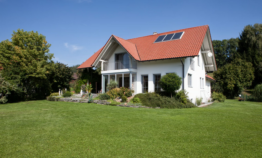 Two-story white fixer-upper house with a red tile roof and large green lawn under a blue sky.